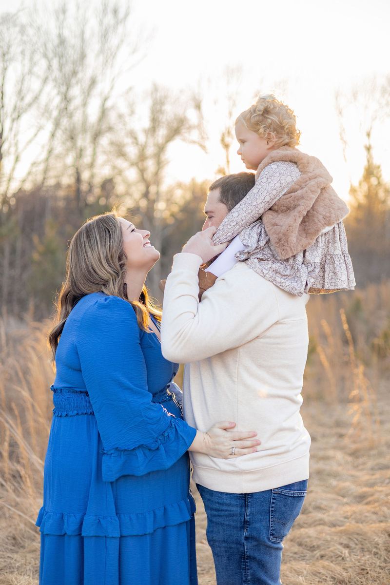 Expecting mom looking up at her toddler on dad's shoulders in golden grass