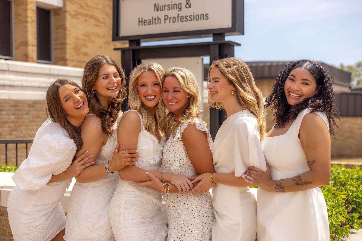 Six graduates in white laughing together outside the nursing building