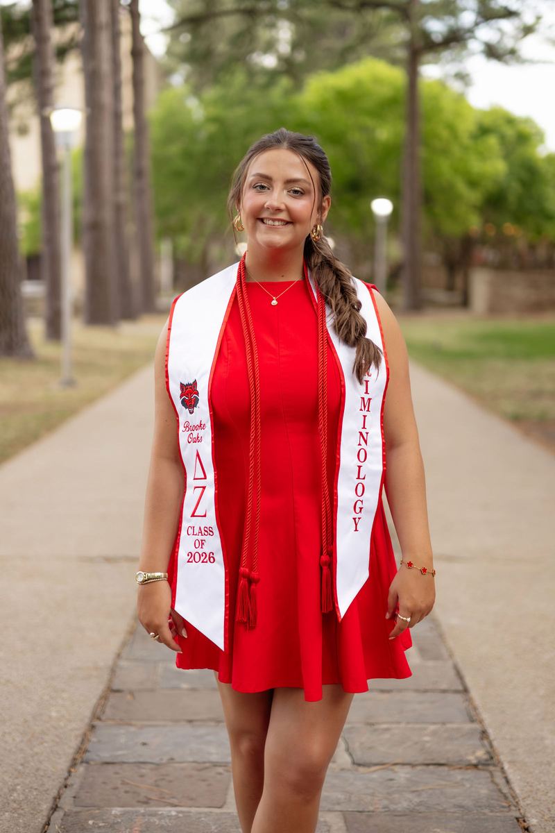 Graduate in a red dress and stole on a tree-lined campus walk
