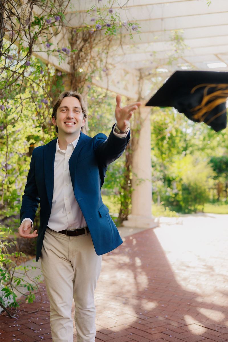 Graduate in a navy blazer tossing his cap under a pergola