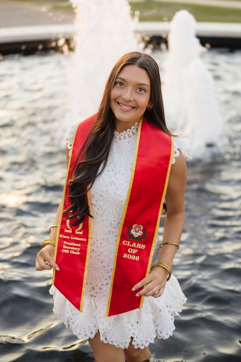 Graduate in a white dress and red stole in front of a fountain