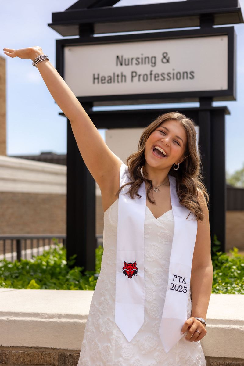 Graduate cheering with one arm up beside the Nursing and Health Professions sign