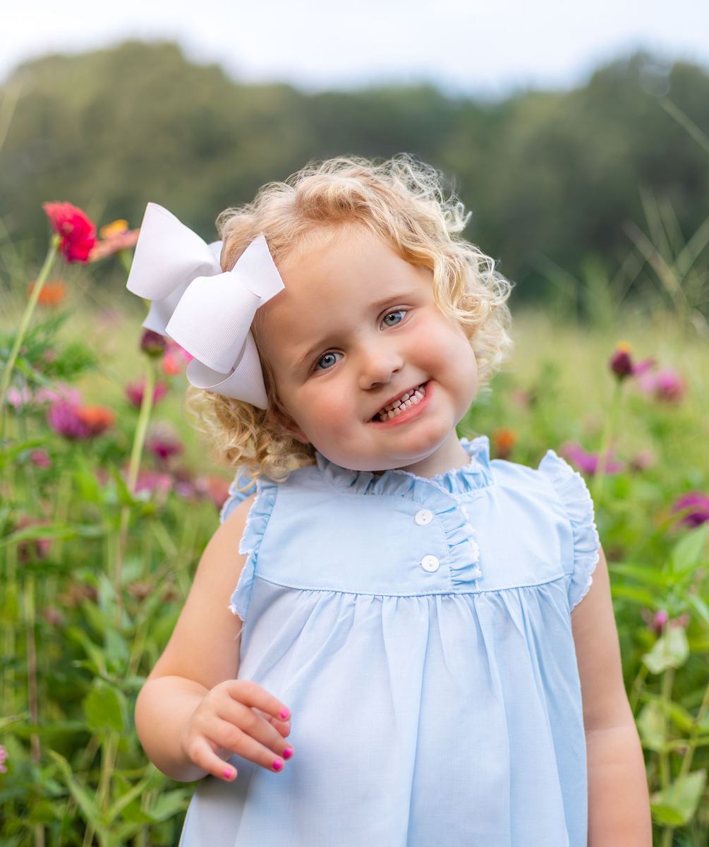 Toddler in a blue dress and white bow smiling among wildflowers